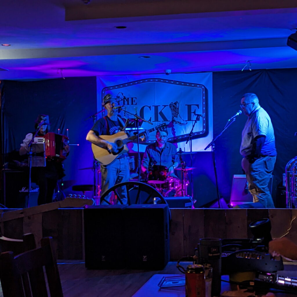 Picture of a gutarist, singer, accordion player, and a drummer performing on stage at The Buckle